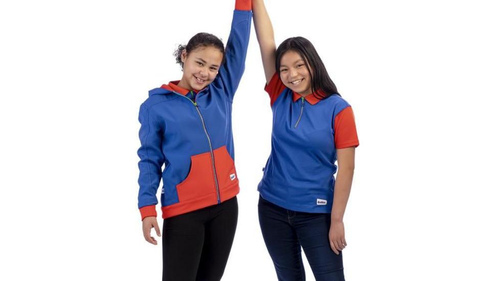 Two girls stand next to one another, they are holding hands in the air. The girl on the left has dark hair tied back and is wearing a blue and red hoodie with a zip down the middle and black leggings. The girl on the right has long, dark hair and is wearing a red and blue polo shirt with a three-quarter zip, and jeans. The background behind them is plain white.