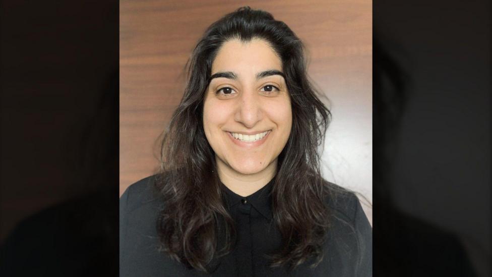 A woman with long dark hair is shown in a portrait against a smooth wooden background. She’s wearing a black collared shirt and looking directly at the camera.