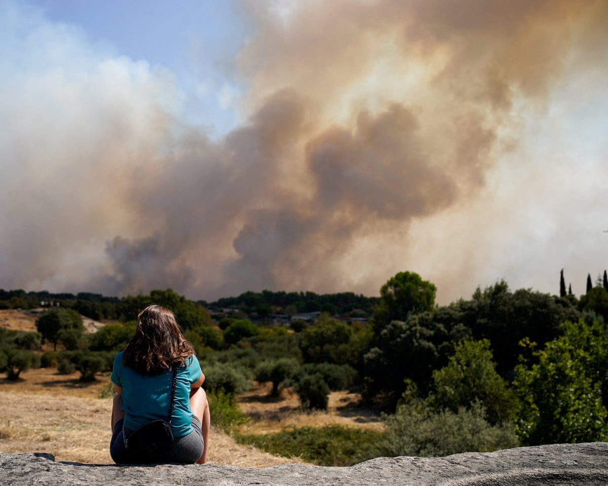 A woman with her back to the camera sits watching a huge cloud of smoke rising from a burning hillside in the distance