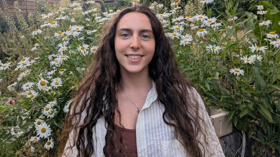 Aviva, a young woman with long dark hair standing in a garden surrounded by white daisies, with greenery in the background.