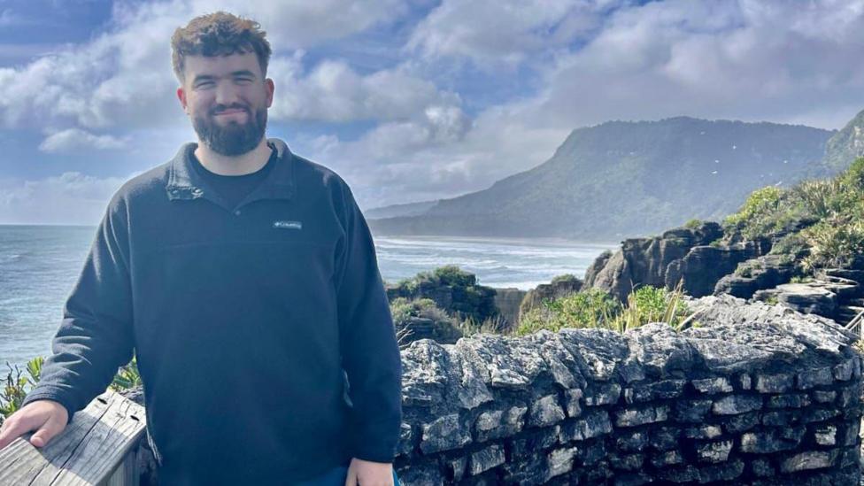 Lloyd, who has dark curly hair and facial hair, is wearing a dark jumper and is standing by a stone wall at a coastal viewpoint, with ocean waves, cliffs, and a cloudy blue sky in the background.