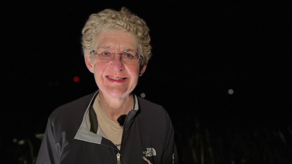 A woman with short, greying hair smiles at the camera. She is wearing glasses and a black zip-up jacket. There is a light on her face but the background is completely dark.