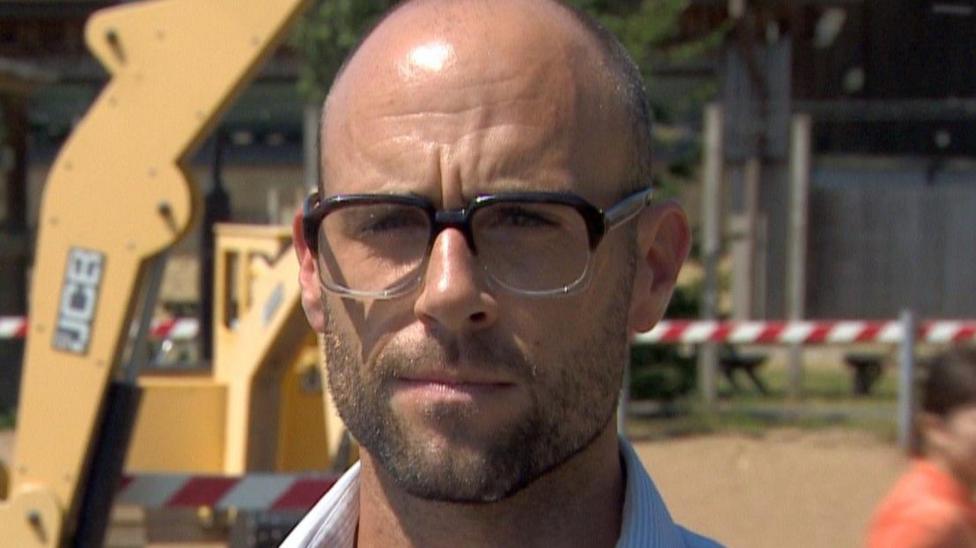 A man is looking at the camera. He has close cropped hair and is wearing glasses.