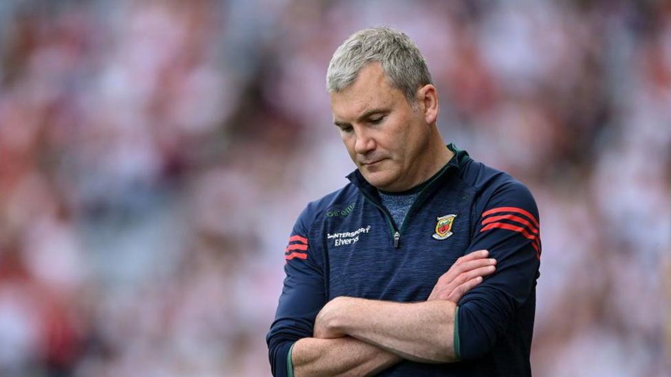 A man with short grey hair looks down at his folder arms. He is wearing a navy and red fleece with a Mayo GAA crest.
