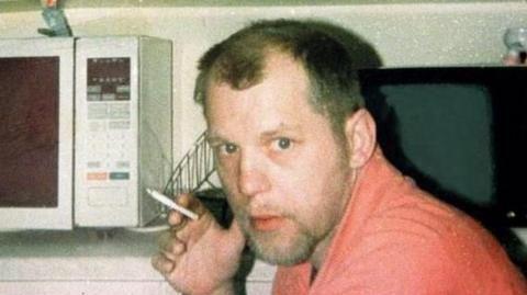 Michael Stone, a man with brown hair and stubble, looking at the camera with a neutral expression. He is wearing a coral coloured top and holds a cigarette in one hand. Behind him is a counter with a television and microwave on it.