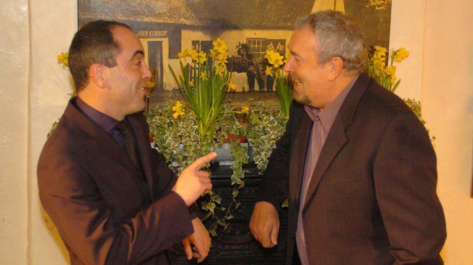 Gerry Kelly and Jimmy Nesbitt talking while both wearing grey blazers and purple coloured shirts.