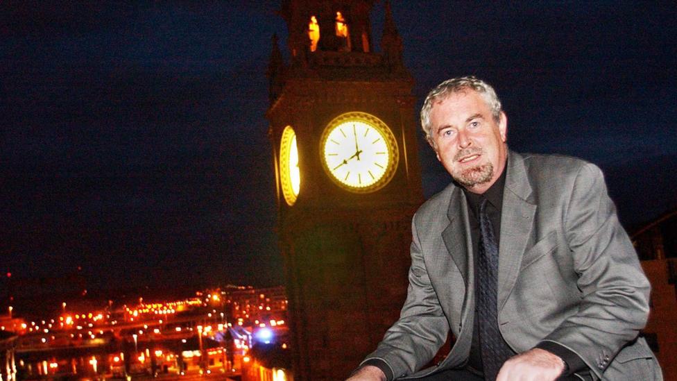 A man with short brown and grey hair wearing a grey suit as he sits in front of a lit up clock tower.