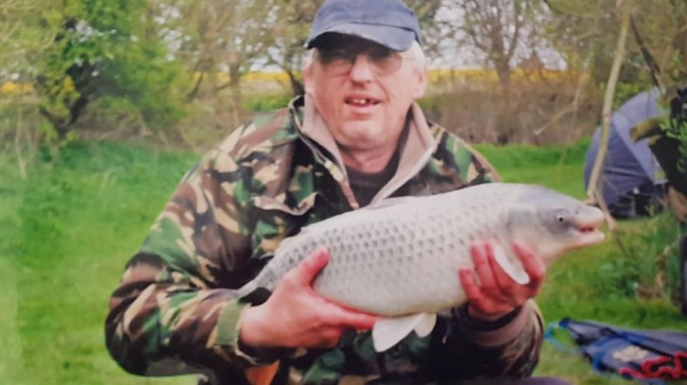 A man looks towards the camera while holding a large white and grey fish that he has caught. He is wearing a camouflage jacket and a blue cap. He is standing on a grassy bank with trees and a blue tent behind him.