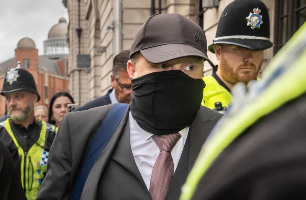 A man wearing a grey suit, white shirt, tie, black cap and face mask, is seen leaving a court building surrounded by police officers wearing hi-vis vests and black uniforms.