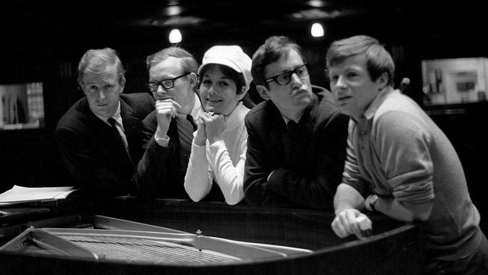 A black and white photo of the cast of I'm Sorry I'll Read That Again in 1965: Tim Brooke-Taylor, Graeme Garden, Jo Kendall, David Hatch & Bill Oddie. They are standing against a large piano.