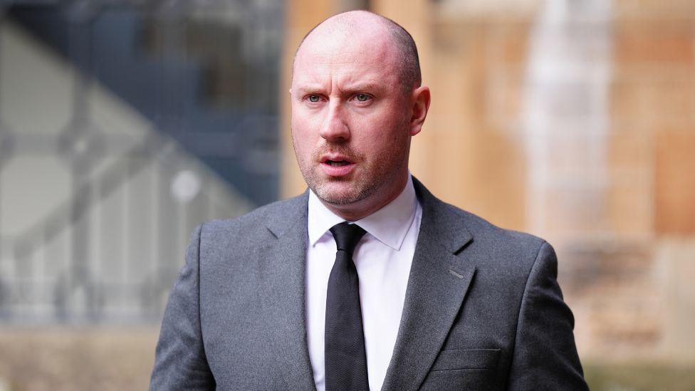 Neil Gray wearing a white shirt with a black tie under a grey suit.