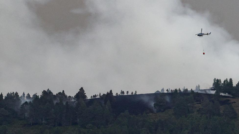 A helicopter in mid-air against a background of a plume of white smoke over a dark landscape.