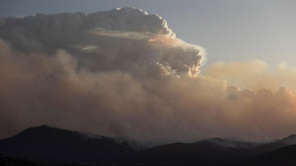 A large cloud above dark hills with smoke surrounding the region