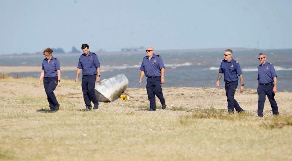 Five people wearing navy shirts and trousers walking in a line along the sand. The sea is behind them and there is a bouquet of yellow sunflowers on the sand.
