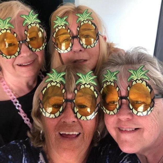 Four smiling older women pose together for a selfie, each wearing oversized novelty sunglasses shaped like pineapples.