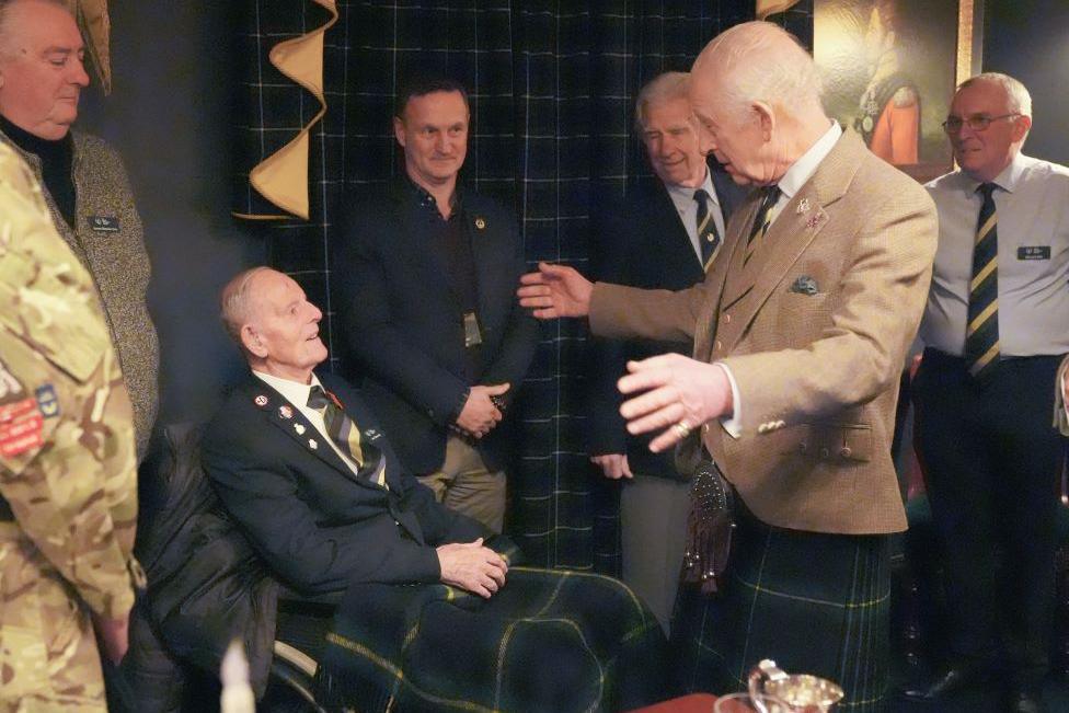 Jim Glennie, 99-year-old man wearing a striped tie and a jacket, speaking to King Charles in a light brown jacket and kilt, with his arms outstretched