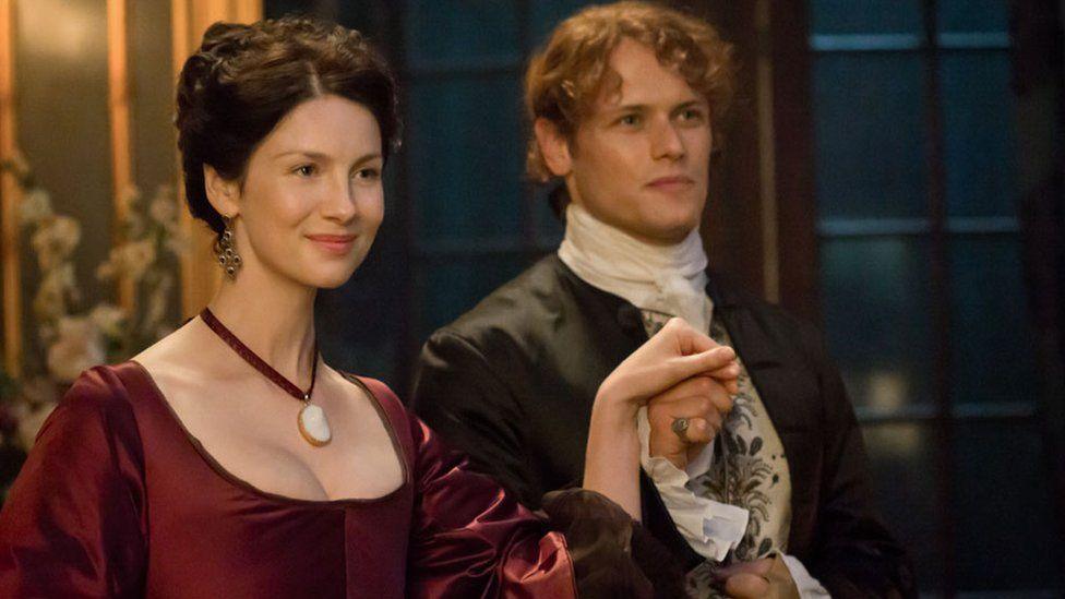 Caitriona Balfe and Sam Heughan in Outlander. They are both in formal 18th Century dress and Sam holds Caitriona's hand in a ballroom. 