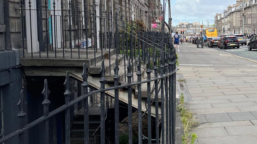 Railings marking the area of a basement. There is a pavement with steps rising up to a green doorway above. there are cars on the street.