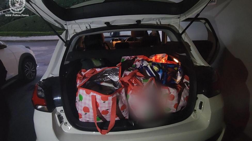 Several red and white bags with apples on them in the boot of a white car.