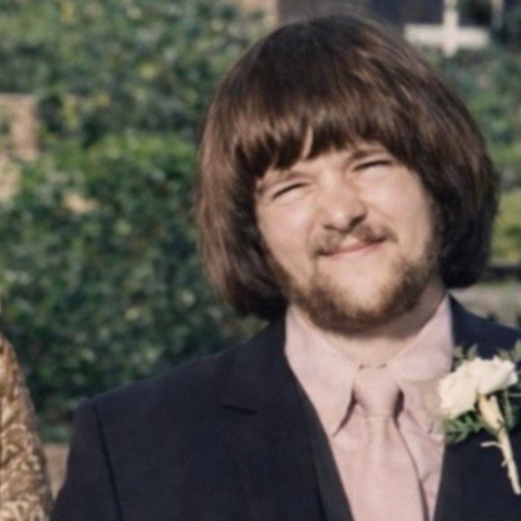 An archive image of Graham Finn with short brown hair and a fringe reaching his eyebrows. He is wearing a dark suit with a pink shirt and pink tie, and a white carnation pinned to his blazer lapel. He is standing in front of a green bush.