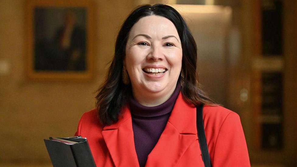 Monica Lennon wearing a bright red blazer over a dark turtleneck top stands indoors holding a black folder. The background is softly blurred, with framed artwork and warm lighting visible.