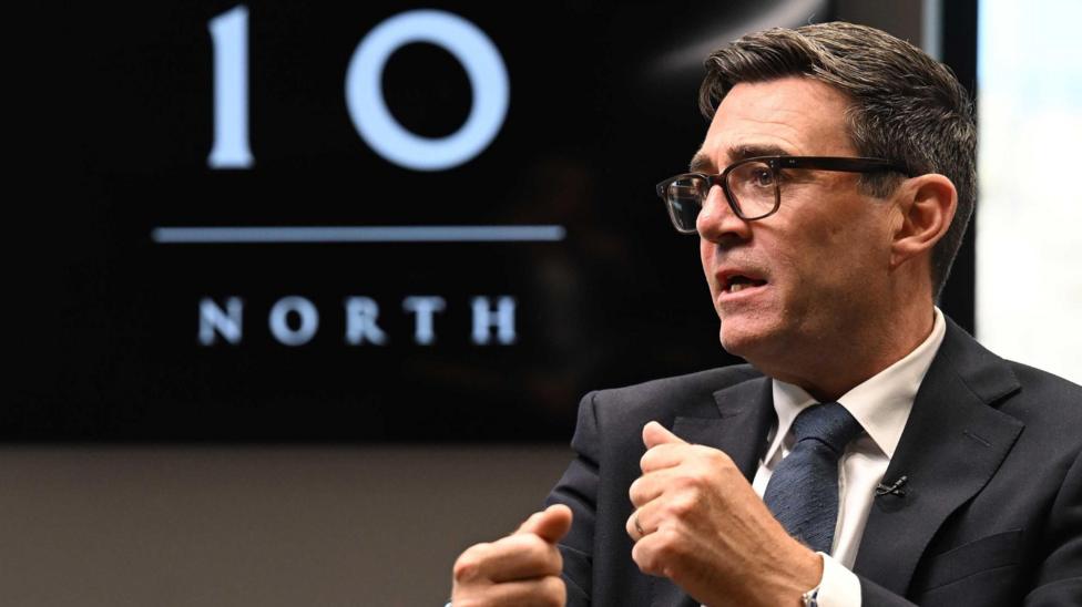 Andy Burnham is pictured in a suit and glasses, gesturing with both hands while seated. A large screen in the background displays the words “10 NORTH” in white text on a black background.