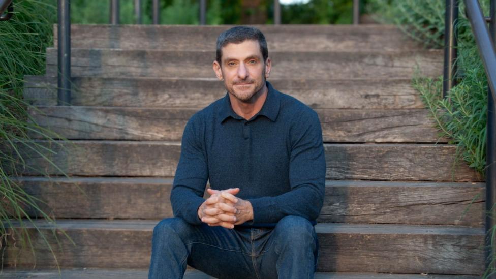 Amir sits on wide wooden outdoor steps, leaning slightly forward with his hands clasped together. He is wearing a dark long‑sleeve top and jeans.