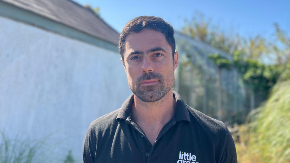 A man with short black hair and stubble looks at the camera, wearing a black polo top with 