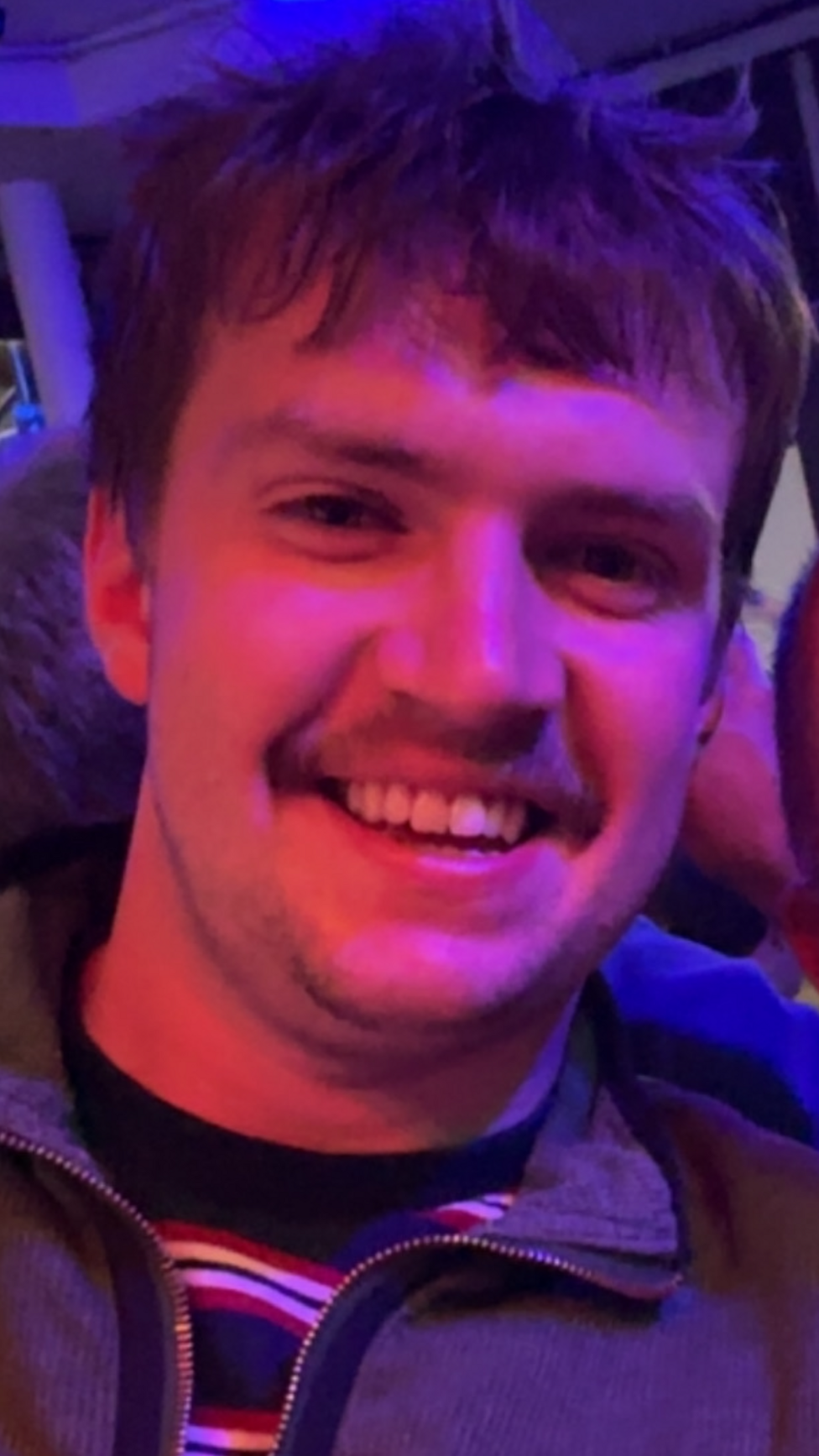 A man smiles at the camera. He has short dark hair and a moustache.