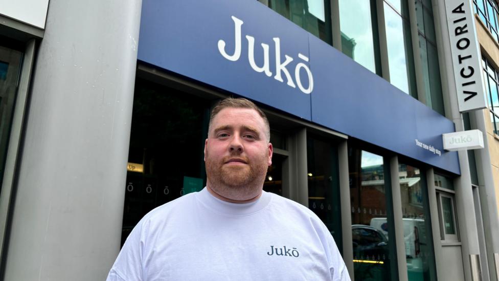 Shane McGann with light short hair and a white top with 'Juko'. He is standing in front of the cafe which also has the shop name on it over a blue background. The building is grey.