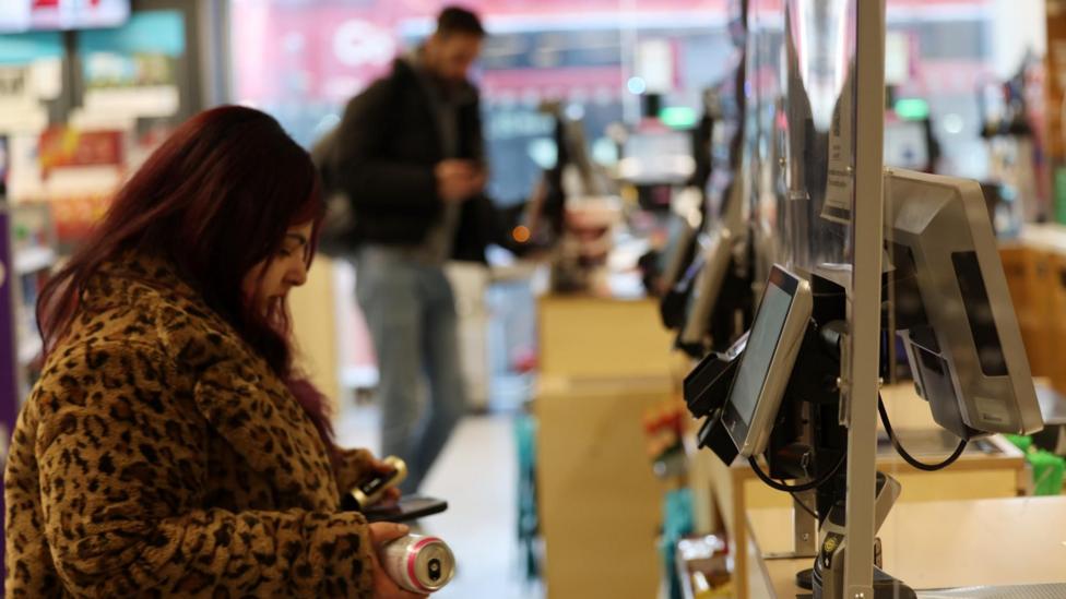 A woman in a patterned coat is standing at a self-checkout in a shop, holding a can of drink.