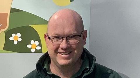 Garry Simmonds is smiling at the camera. He is balding and is wearing spectacles and a green top.