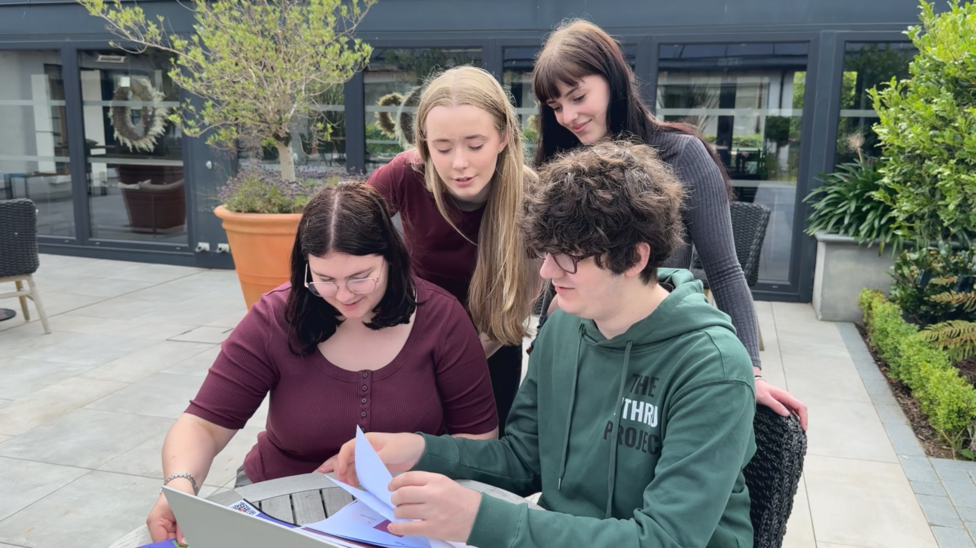 Four young people, two standing and two sitting, are looking at a laptop and some paper.