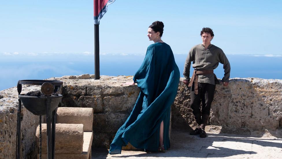 Anne Hathaway as Penelope (Odysseus's wife) in centre of image in blue gown looking to the side and Tom Holland as their son Telemachus leaning on wall in khaki top and dark trousers.