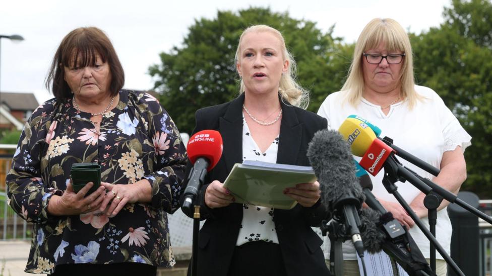 Three women, one with shoulder length brown hair and a fringe, another in a black suit with long blonde hair tied up, and a third with long blonde hair with a fringe, standing beside each other in front of media microphones. 
