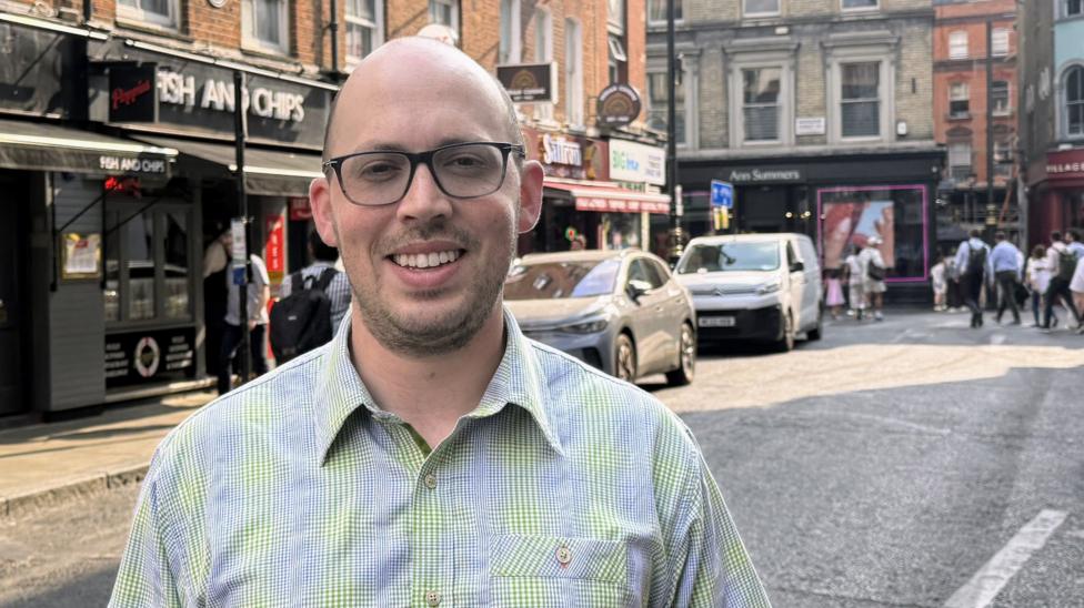 Matt Cohen stands in a street in London's Soho, wearing glasses and checked shirt