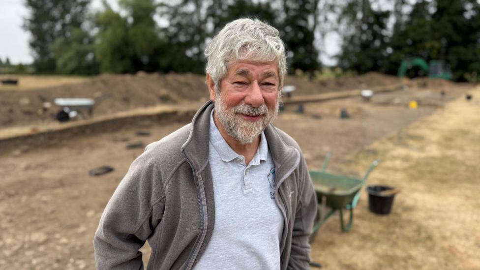 Dr Peter Halkon pictured standing next to an area that has ben excavated. He has grey hair and a grey beard and is wearing a brown fleece and grey polo shirt.