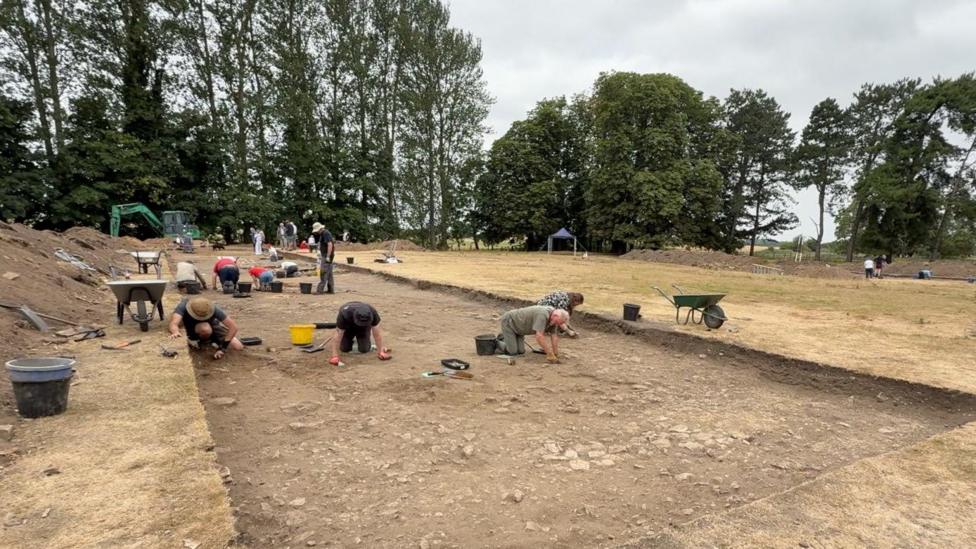 A group of people on their hands and knees on a large plot of soil. They are picking up bits of Roman material that has been discovered underneath the ground.