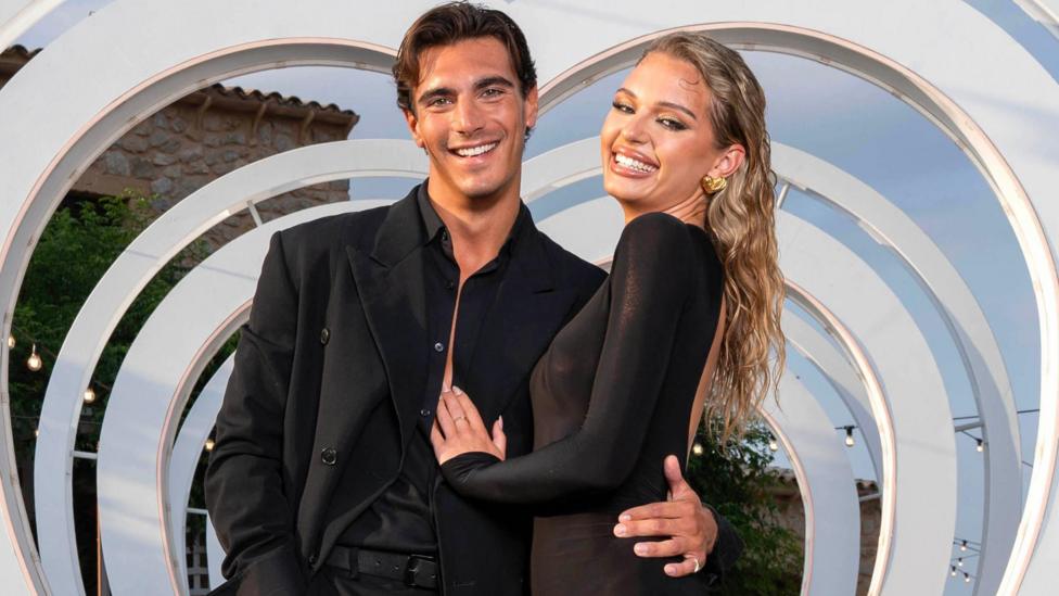 Lorenzo Alessi and Julia Majchrzak pose in front of a heart-shaped cut-out. Lorenzo is wearing a black suit and Julia a black dress. 