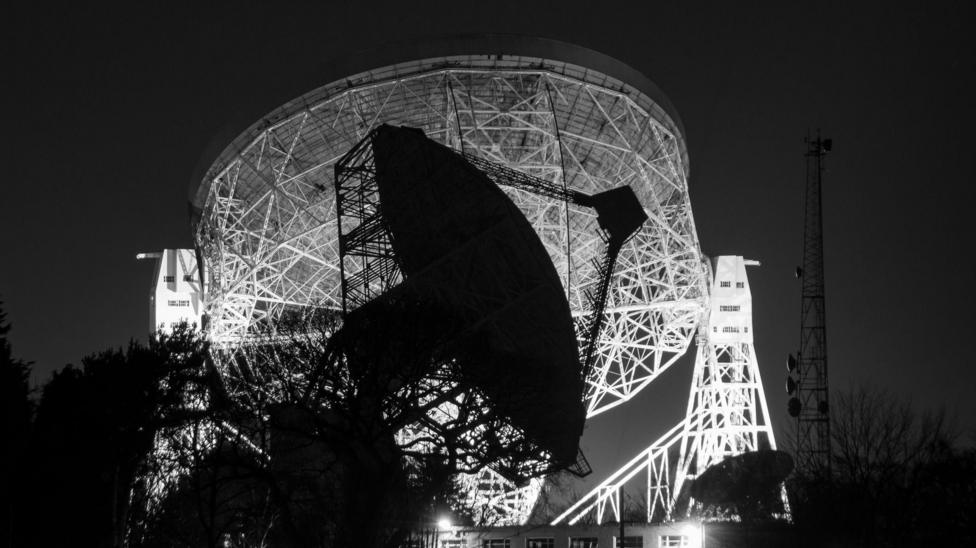 The Mark II telescope seen illuminated at night behind the Lovell Radio Telescope at Jodrell Bank Observatory.