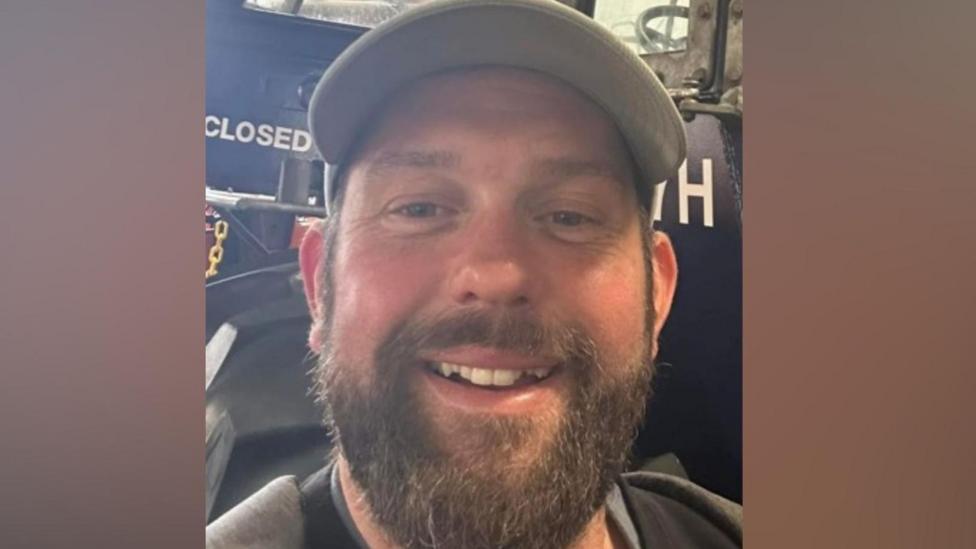 James Desborough is smiling. He has a brown beard and is standing in front of a blue tractor. He has a grey baseball cap on.