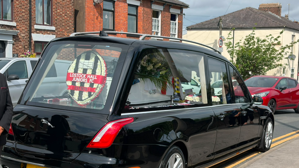 A white coffin covered with white flowers and a wreath of the Lostock Hall Junior Football Club logo are in the rear window of the black hearse at Kerry Coombes funeral.