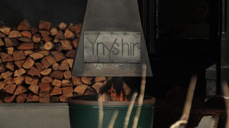 The sign of Ynshir restaurant's name engraved on a fire pit. There's a fire burning with logs stacked up in the background. 