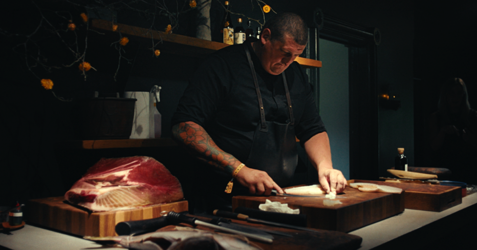Ward cutting fish on a brown wooden chopping board. He is wearing a black top and a dark apron on top and has a sleeve tattoo on his right arm. There is a slab of red meat on the side too. 