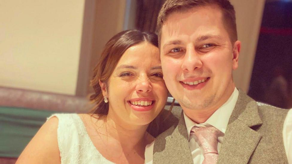 A wedding photo of Josh and Kelly. Kelly wears a white, embroidered sleeveless wedding dress and pearl earrings and her hair is in an up-do. Josh wears a white shirt, pale pink embroidered tie and beige waistcoat. They are both looking at the camera and smiling.