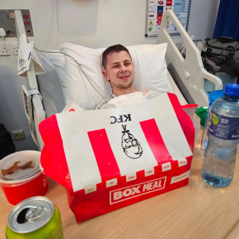 Josh wearing a hospital gown and lying in a hospital bed with white bedding. In front of him is a tray with a bottle of water, a red and white cardboard KFC box, a KFC gravy pot and an open can of pop on it.