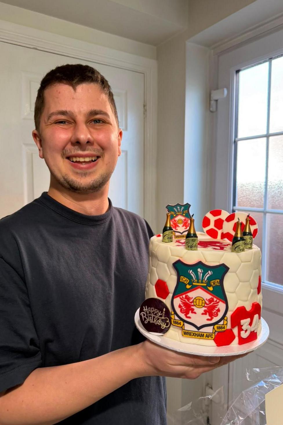 Josh wearing a dark t shirt and holding an birthday cake designed with Wrexham FC branding. He is smiling and looking at the camera.