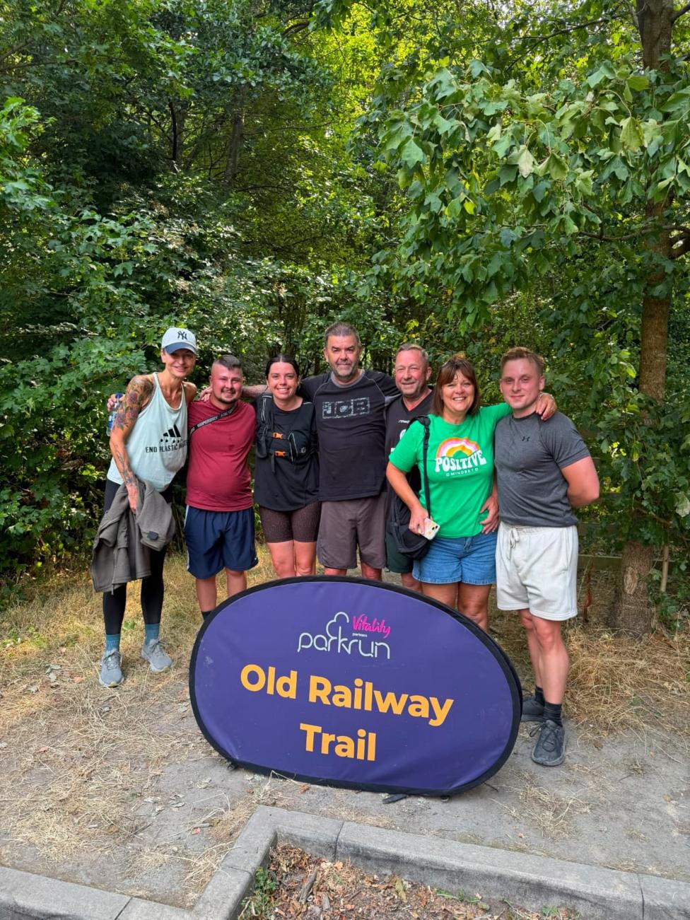 Seven people stood behind a parkrun-branded purple and orange sign which reads: Old Railway Trail. They have their arms around each other and are smiling and looking at the camera. Behind them are trees and bushes.