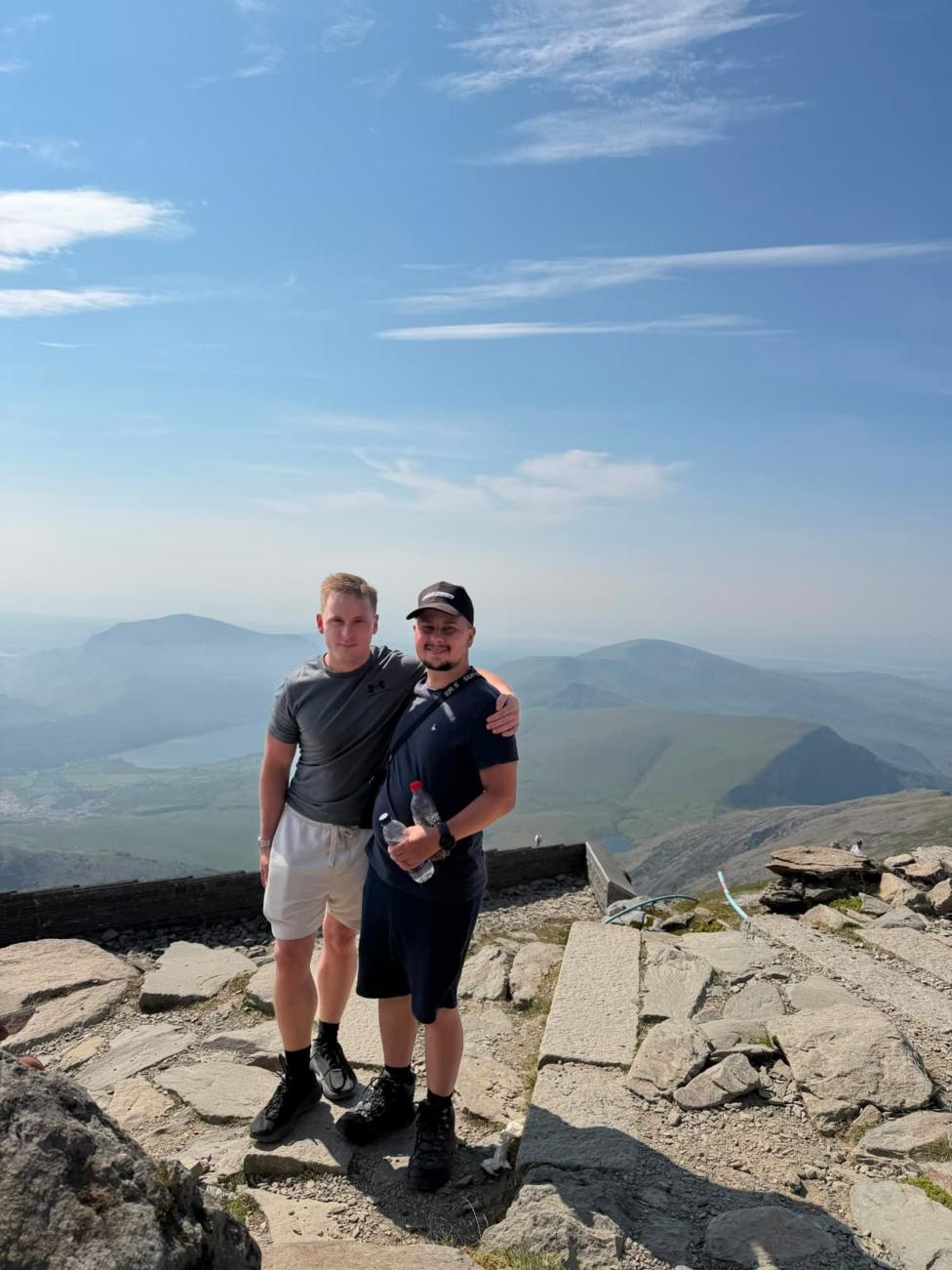 Two men, one blonde, the other brunette and wearing a cap. They are both wearinf t shirts, shorts and hiking boots and have an arm around each other. The man with the cap is holding two plastic water bottles. They are stood at the top of a mountain with hills and sky as a backdrop. They are looking at the camera.