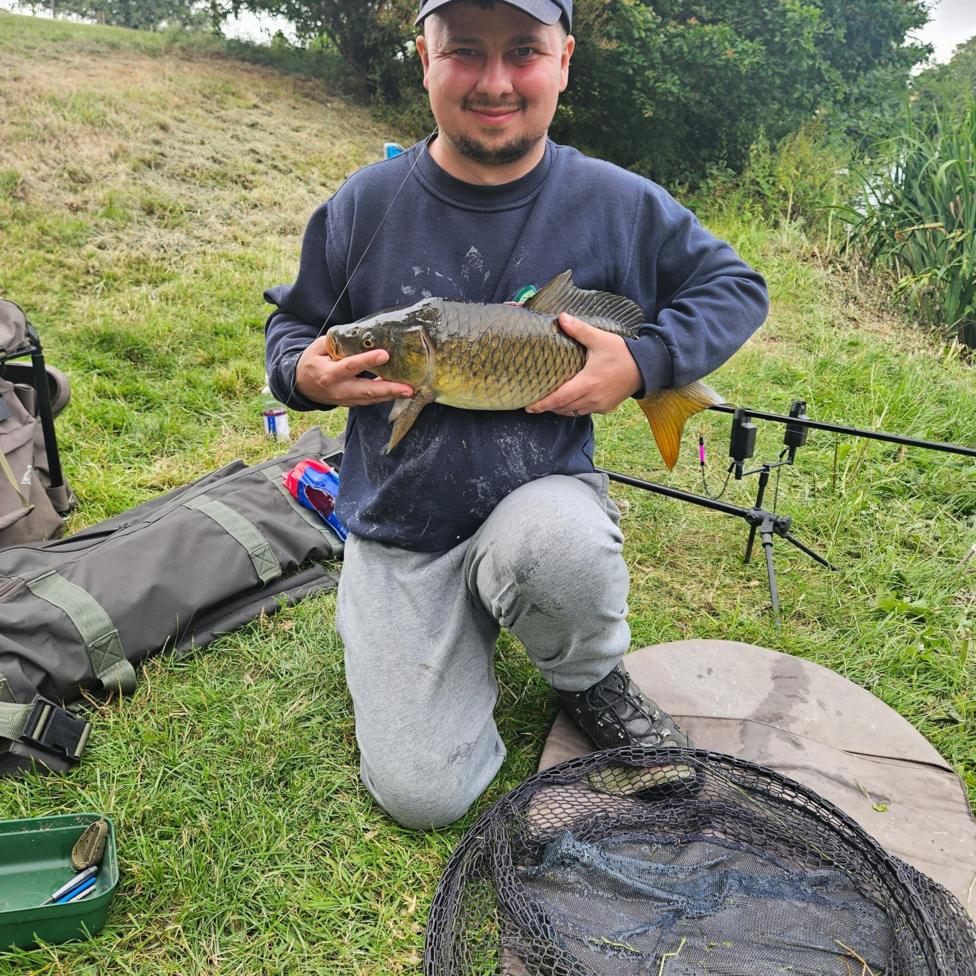 A man wearing a navy cap, navy sweatshirt and grey tracksuit bottoms nealing down on a patch of grass and holding a large, scaley fish in his hands. He is looking at the camera and smiling. Around him on the grass are various pieces of fishing equipment. 